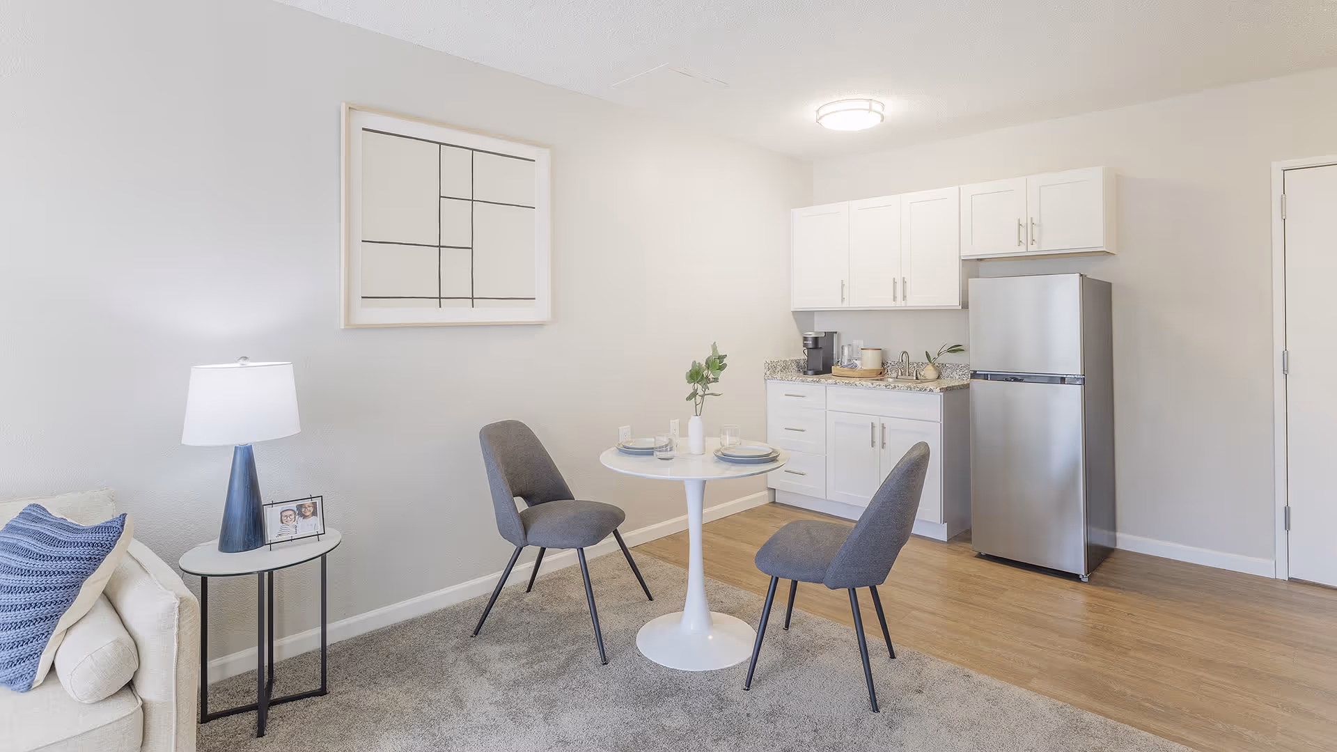 A small dining area with a round white table set for two, two gray chairs, a kitchenette with white cabinets, a stainless steel refrigerator, and a small sink. To the left, there is a beige couch with a blue pillow and a side table with a lamp and a framed photo. The room has light-colored walls and a mix of carpet and wood flooring.