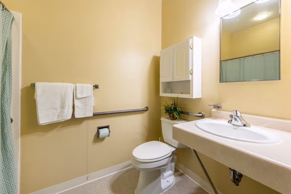 A clean bathroom with beige walls featuring a white toilet, a white sink with a silver faucet, a wall-mounted cabinet above the toilet, a mirror above the sink, a towel rack with two white towels, and a shower curtain with a green and white circular pattern.