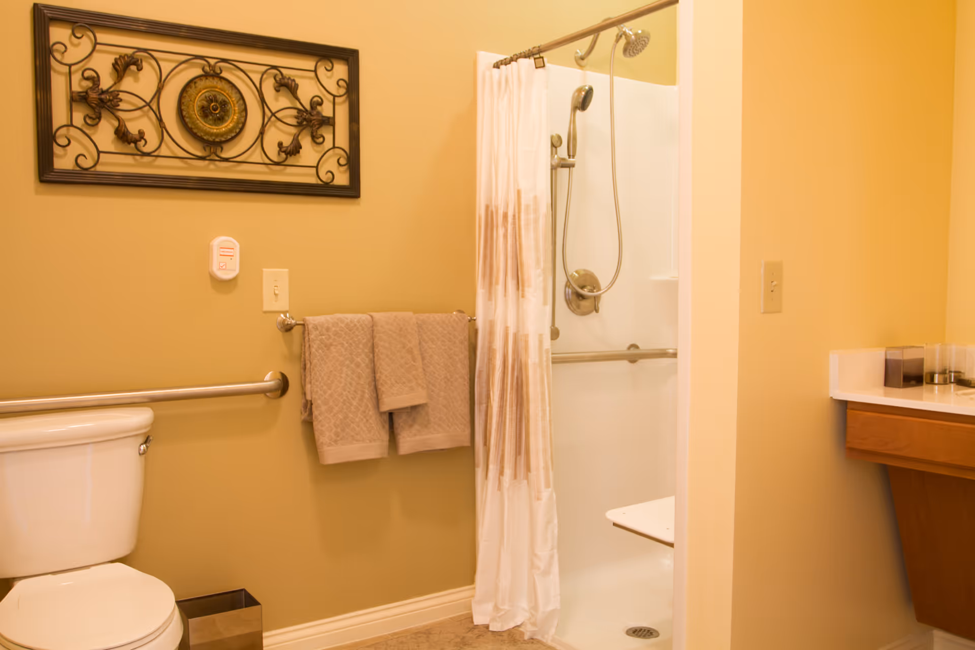 A bathroom with beige walls featuring a white toilet with a metal grab bar beside it, two beige towels hanging on a towel rack, a shower with a white curtain and a handheld showerhead, and a wooden vanity with a white countertop.