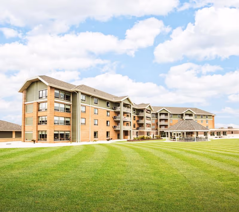 Four-story senior living building with balconies, a gazebo, and a large manicured lawn under a partly cloudy sky.