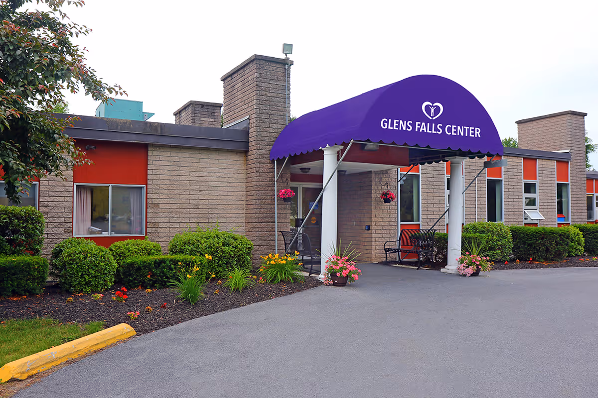 Exterior view of Glens Falls Center showing the entrance with a purple awning displaying the facility's name and logo. The building is made of light brown bricks with red window frames, surrounded by neatly trimmed bushes and flower beds. There is a paved driveway leading to the entrance.