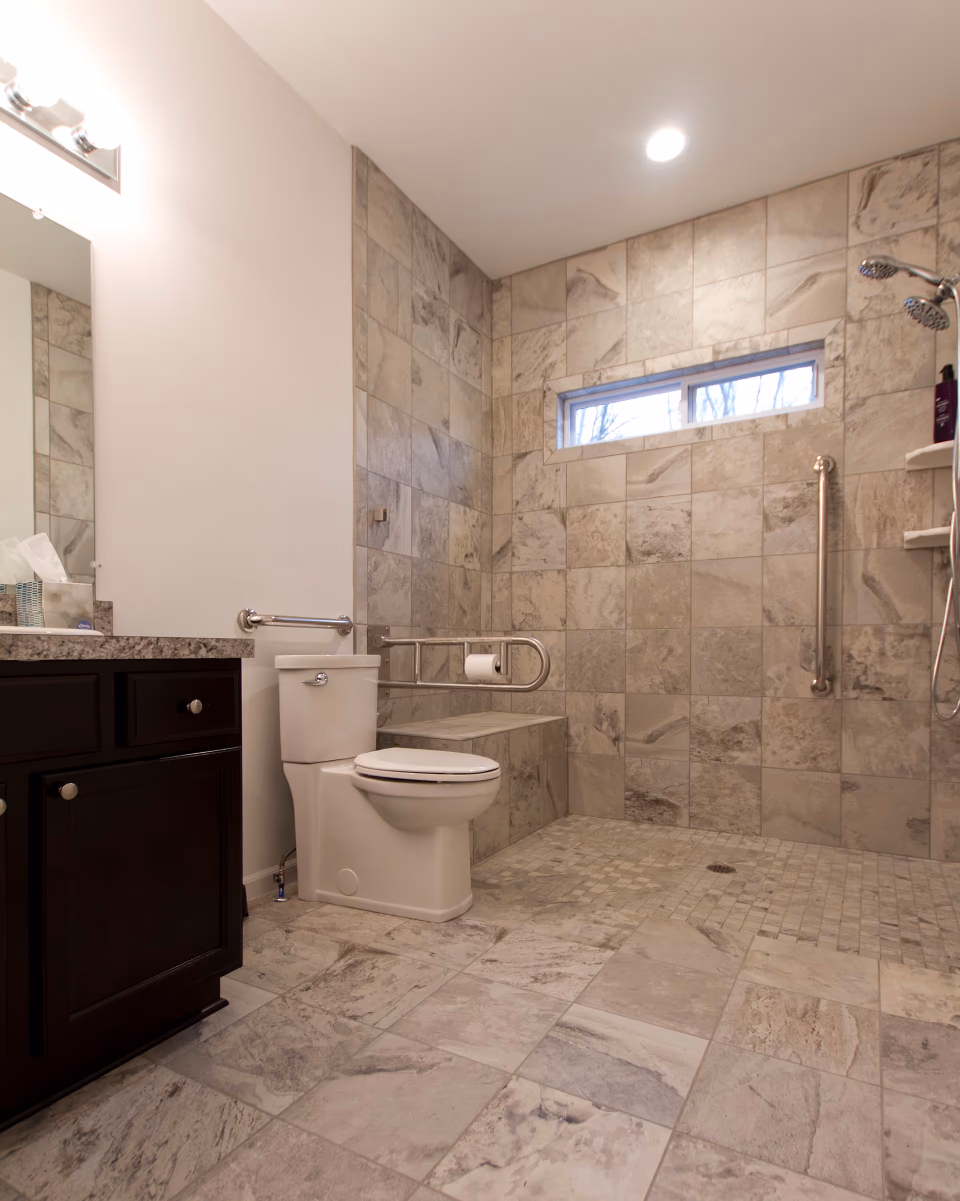 A spacious bathroom with beige and gray tiled walls and floor. It features a white toilet with a metal grab bar and a built-in bench next to it. There is a walk-in shower area with two showerheads and a vertical grab bar. A dark wood vanity with a granite countertop and a mirror with lights above is visible on the left side.