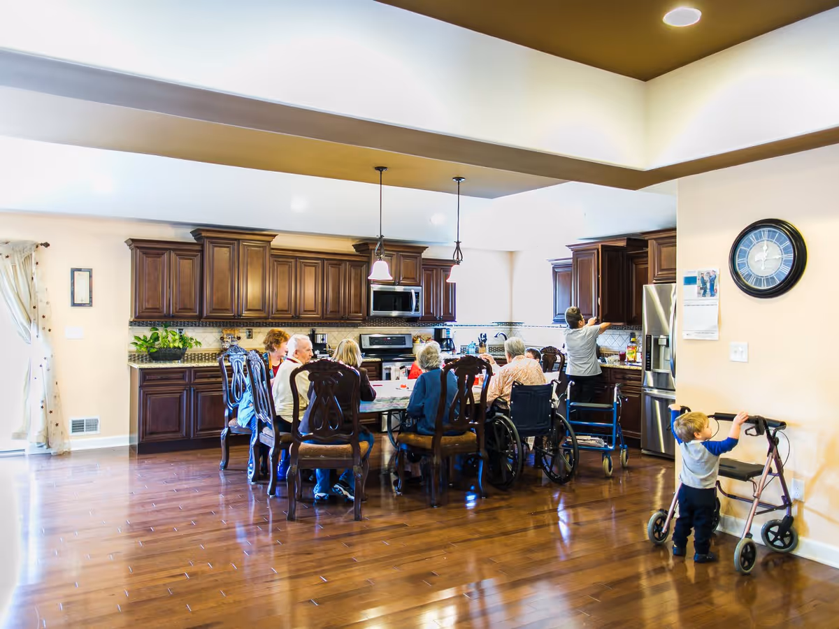 Several elderly residents seated around a dining table in a large open kitchen/dining area while a small child stands by a walker nearby.