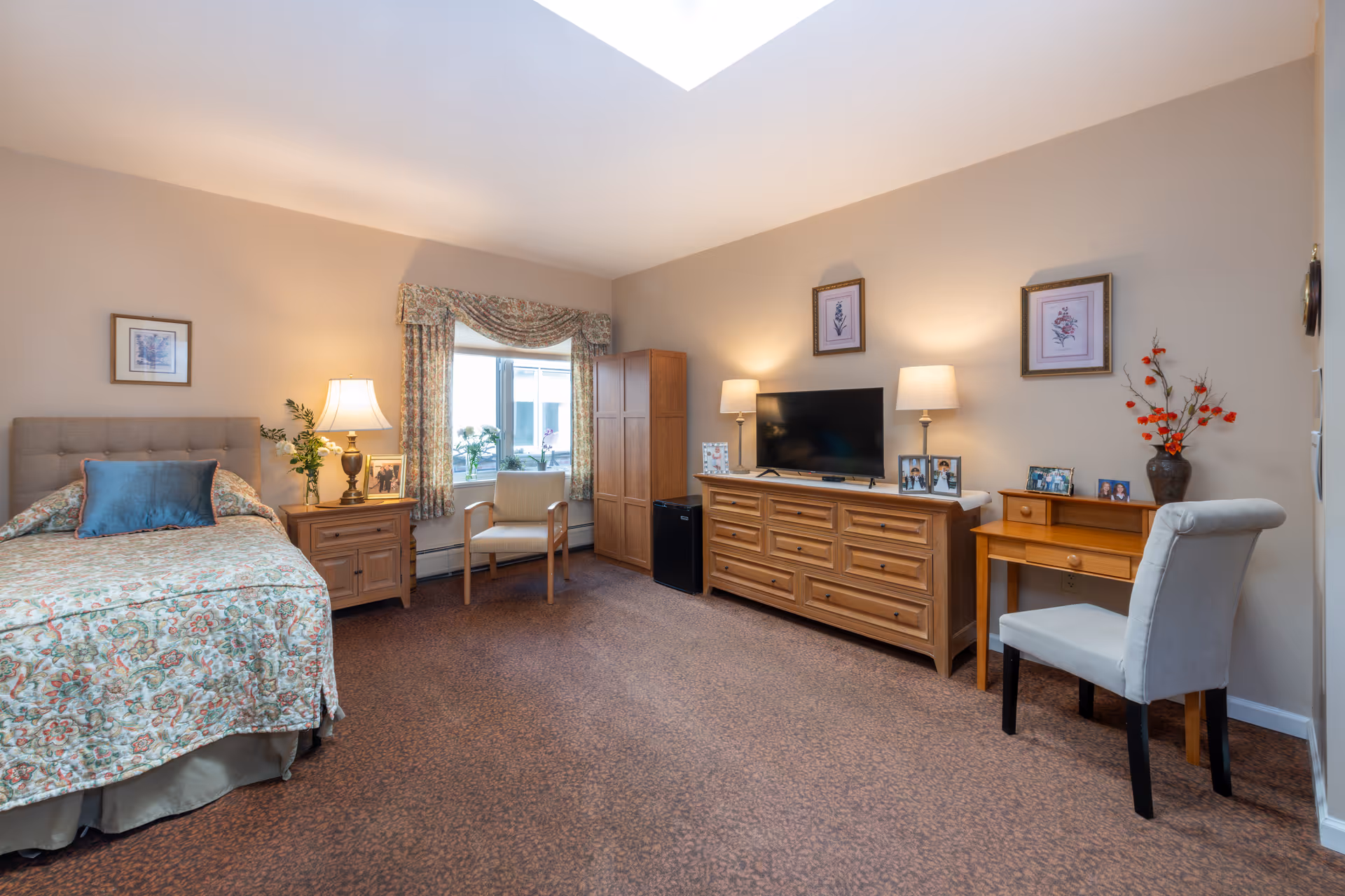 Bright furnished senior residence room with a bed, dresser and TV, seating area, and desk.