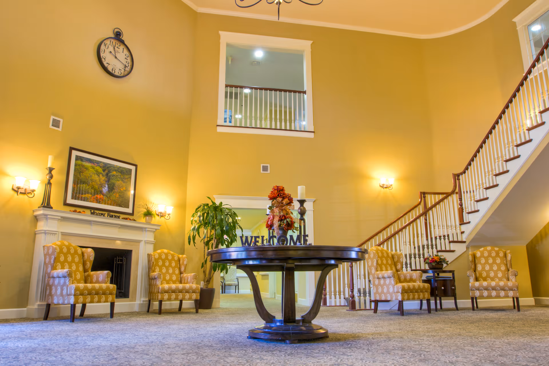 A spacious and warmly lit living room area with high ceilings, featuring a round wooden table in the center decorated with a floral arrangement and a 'WELCOME' sign. Surrounding the table are four patterned yellow armchairs, a fireplace with a framed landscape picture above it, a green potted plant, and a staircase with white railings leading to an upper floor.