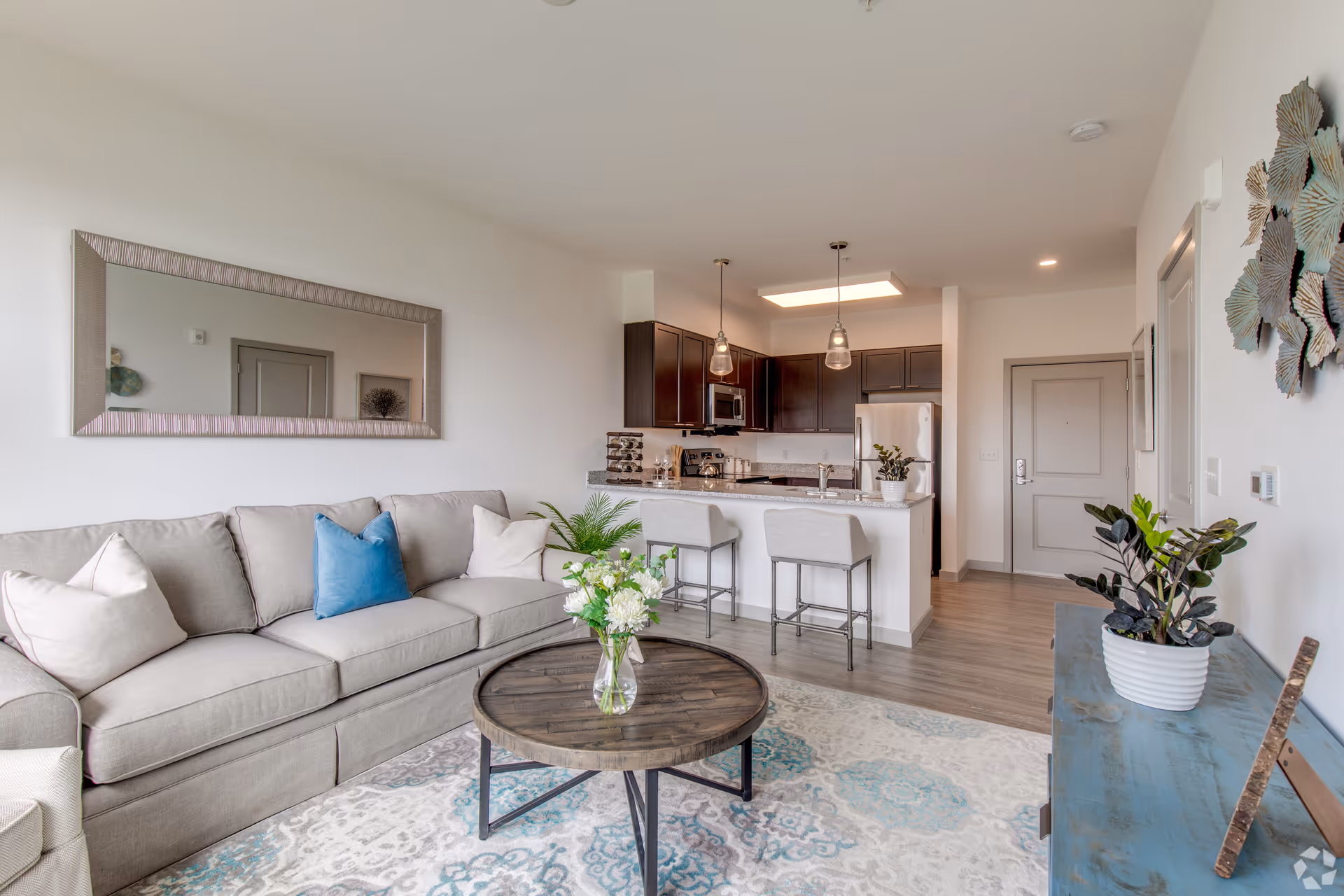 A modern living room and kitchen area with a beige sofa adorned with white and blue pillows, a round wooden coffee table with a vase of flowers, a large wall mirror above the sofa, and a kitchen with dark wood cabinets, stainless steel appliances, and two bar stools at the counter. The room has light-colored walls, a patterned rug, and decorative plants on a blue console table and kitchen counter.