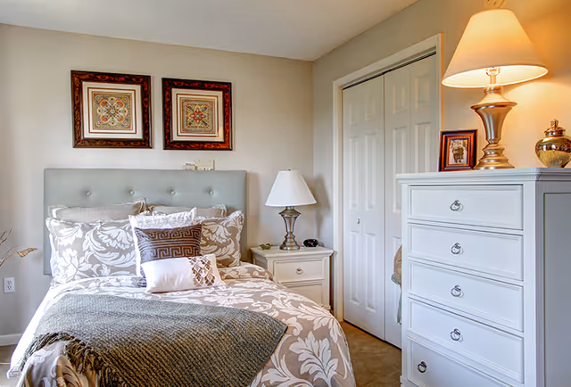 A neatly made bedroom with a patterned bed and pillows, a nightstand and lamp, a white chest of drawers, and closed closet doors.