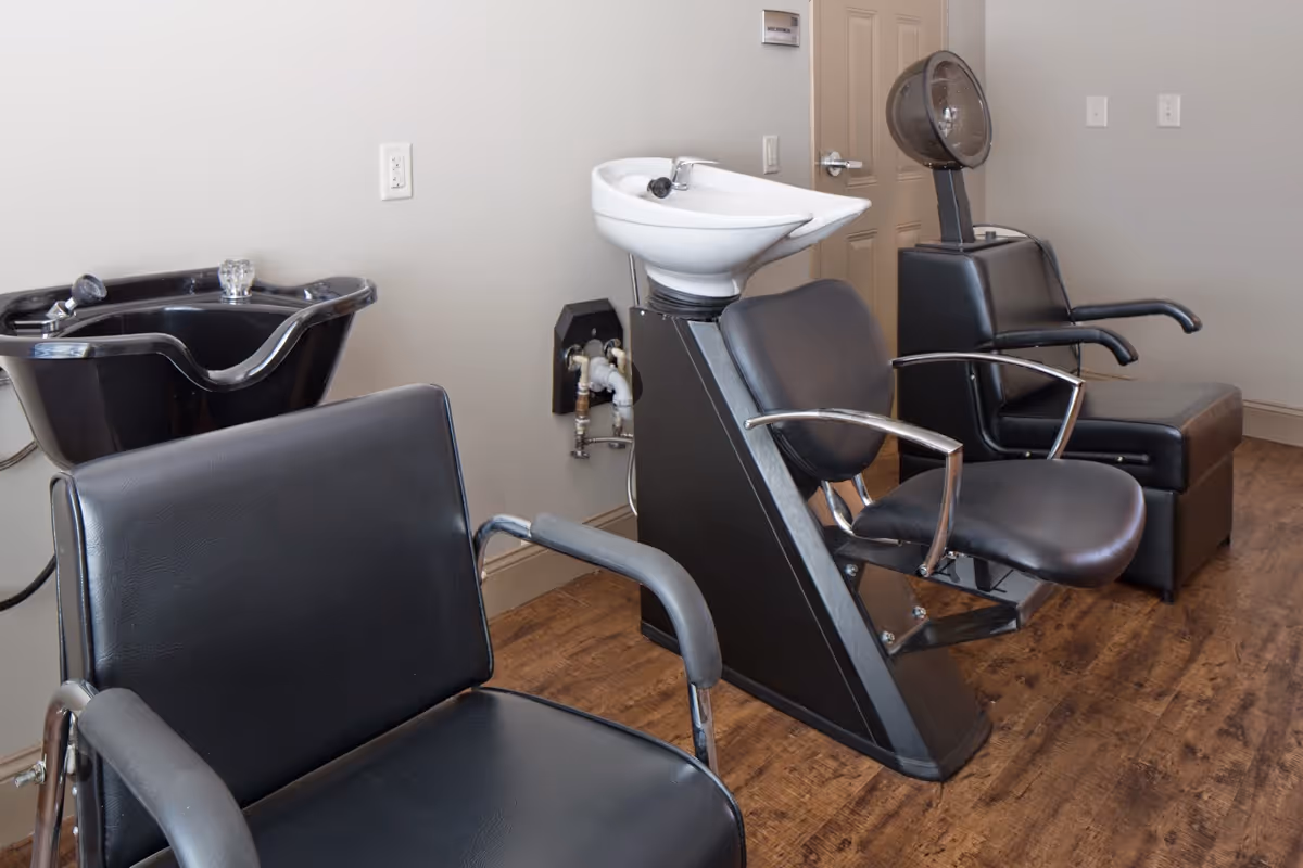 Interior view of a senior living facility's hair salon area featuring black salon chairs, a white shampoo basin, and a hair dryer chair against a beige wall and wooden floor.