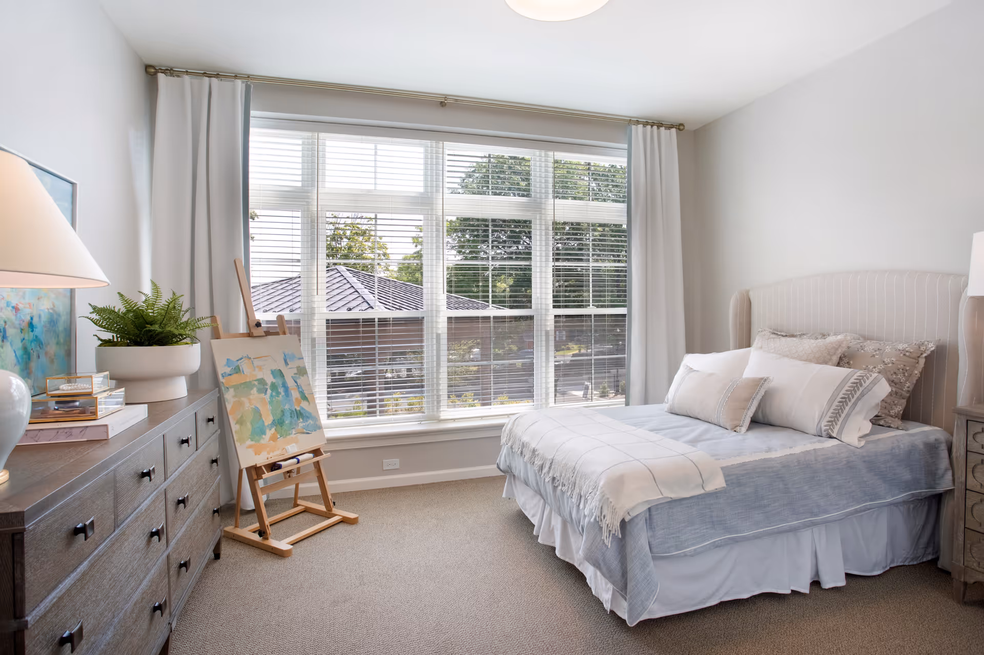 A bright bedroom with a large window covered by white blinds and curtains. The room features a bed with multiple pillows and a light blue blanket, a wooden dresser with a lamp and a potted plant, and an easel holding a colorful painting. The walls are painted light gray and the floor is carpeted.