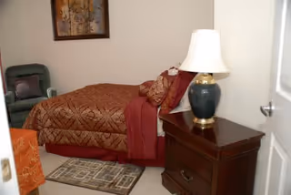 A small bedroom with a bed covered in a patterned brown and red bedspread, a wooden nightstand with a dark green lamp with a white lampshade, a green armchair in the corner, a framed artwork on the wall, and a small patterned rug on the floor.