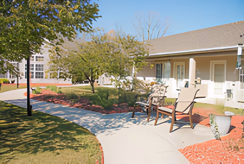 Outdoor patio area at The Wyndmoor of Portage featuring a curved concrete walkway, two chairs, a small table, trees, and landscaped garden beds with red mulch. A building with a covered porch and multiple doors is visible in the background.