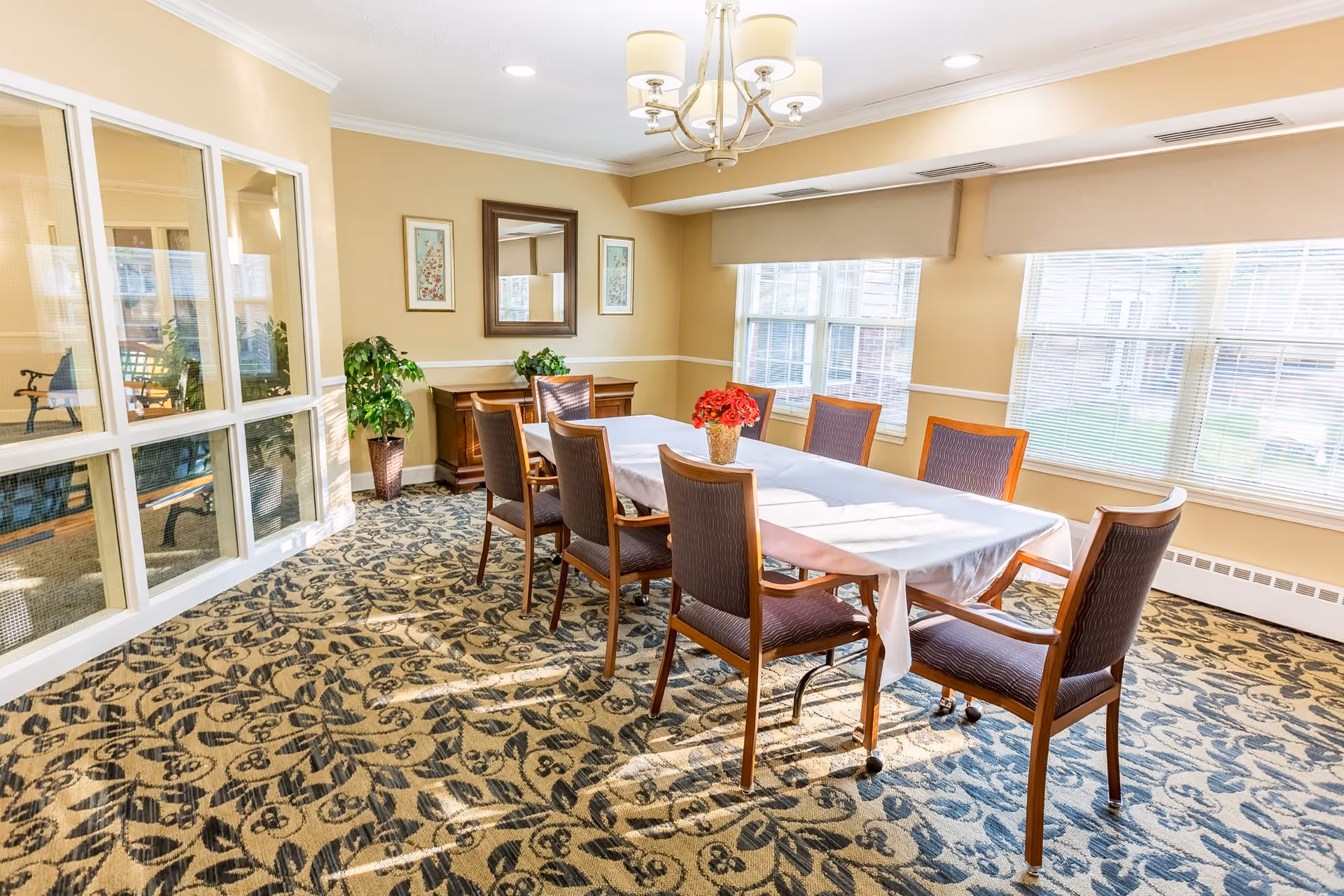 A bright dining room with a rectangular table covered with a white tablecloth and surrounded by eight wooden chairs with cushioned seats. A vase with red flowers is placed in the center of the table. The room has large windows with blinds allowing natural light to fill the space, beige walls, a patterned carpet, a chandelier, and decorative plants and framed artwork on the walls.
