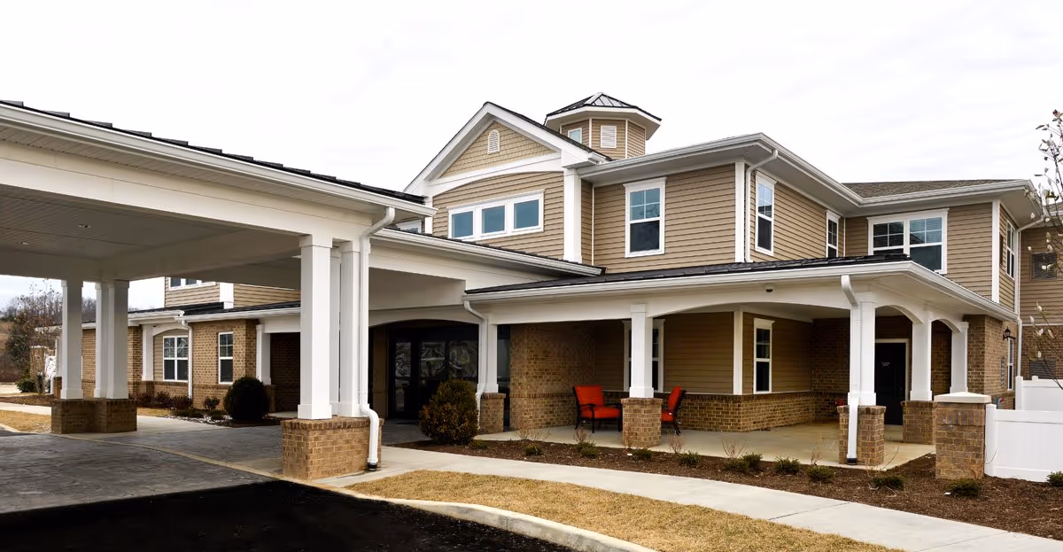 Exterior view of a two-story senior living facility building with beige siding and brick accents. The entrance features a covered drop-off area supported by white columns. There are two red chairs on the porch near the entrance, and a paved walkway leads to the door. The sky is overcast.