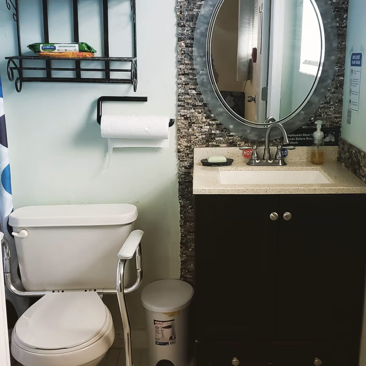 A bathroom with a white toilet equipped with safety rails, a toilet paper holder with a roll of paper, a small trash can, a sink with a beige countertop and dark cabinet, an oval mirror with a decorative frame, a soap dish with a bar of soap, and a hand soap dispenser. There is a tiled backsplash behind the sink and mirror.