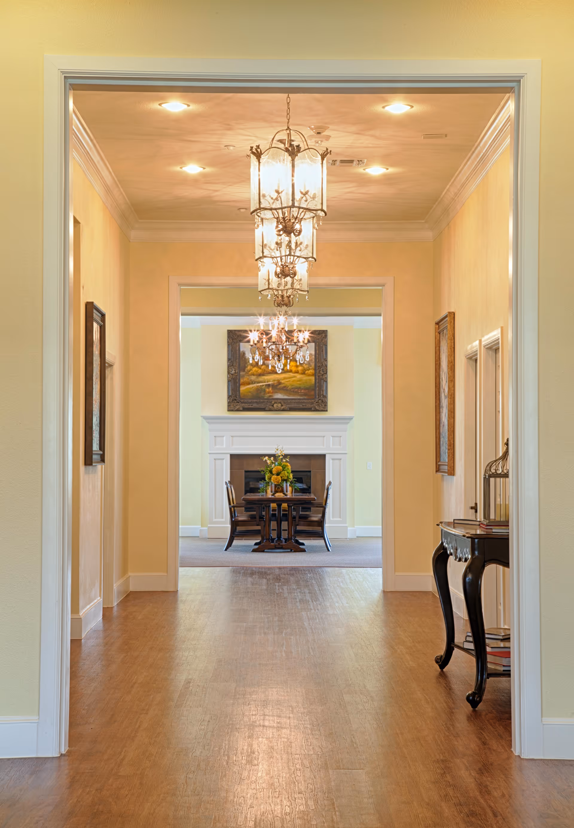 View down a hallway with wooden flooring leading to a dining area with a table and chairs in front of a white fireplace. The hallway has yellow walls, framed artwork on the sides, and two ornate hanging light fixtures on the ceiling. A small black side table with decorative items is on the right side of the hallway.