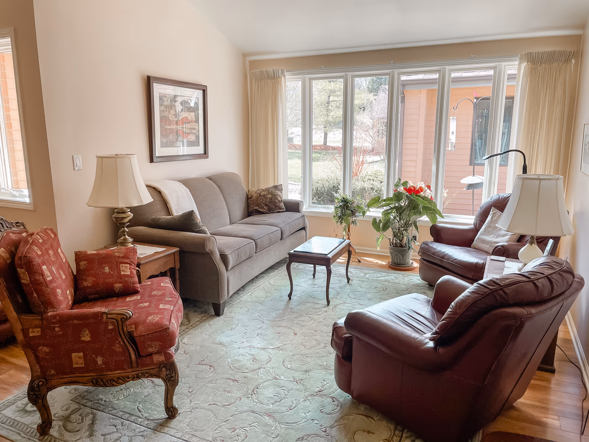 Well-lit living room with a gray sofa, red upholstered and leather armchairs, a small coffee table and large windows with potted plants.