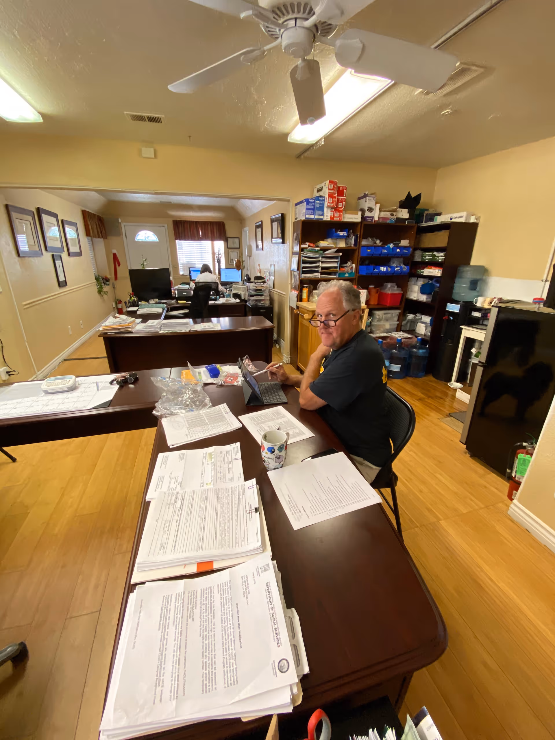 An office space with two desks arranged in an L shape. A man wearing glasses and a black shirt is seated at one desk, looking at a tablet. The desks are covered with papers, documents, and a coffee mug. In the background, another person is seated at a desk working on a computer. Shelves filled with office supplies and water bottles are visible against the wall. The room has wooden flooring, beige walls, and a ceiling fan with lights.