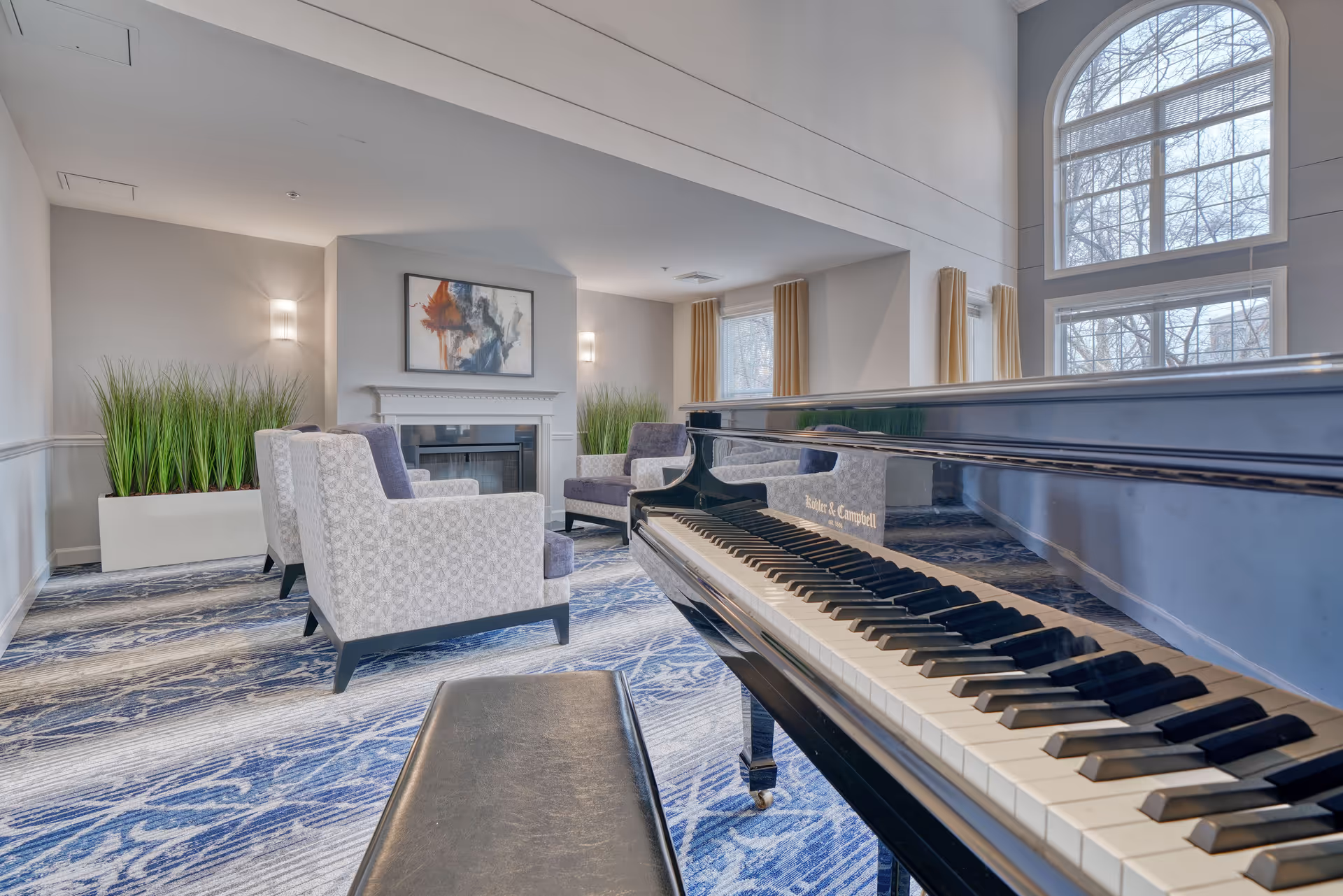 A bright and spacious living room with a grand piano in the foreground, upholstered armchairs arranged around a fireplace, large windows with beige curtains, and decorative plants along the walls.