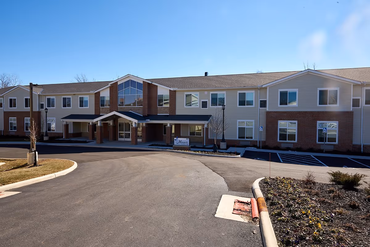 Front exterior view of Monarch Assisted Living facility showing a two-story building with beige siding and brick accents, multiple windows, a covered entrance, a driveway, and a landscaped area with small plants and trees under a clear blue sky.