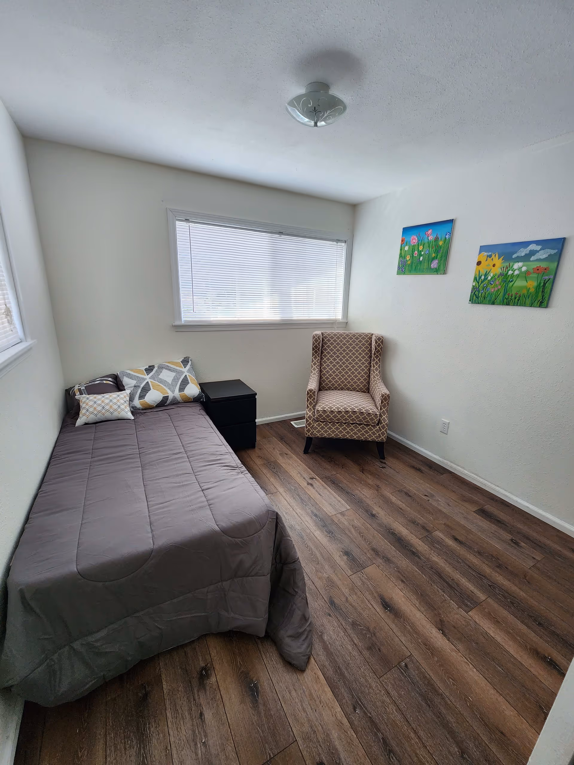 A small bedroom with a single bed covered with a gray comforter and three decorative pillows. Next to the bed is a black nightstand. Across from the bed is a patterned armchair. The room has wood flooring, white walls, a window with blinds, and two colorful paintings of flowers on the wall.