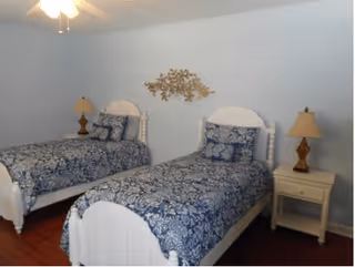 A bedroom with two white twin beds featuring blue and white patterned bedding. Each bed has matching pillows and is accompanied by a wooden nightstand with a lamp. The walls are painted light blue, and there is a decorative metal wall art piece above the beds. The floor is wooden.