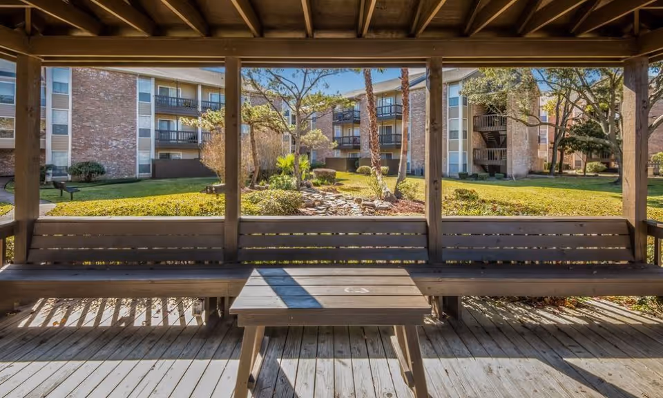 Covered wooden gazebo with built-in benches and a small table overlooking a landscaped courtyard and multi-story apartment buildings.