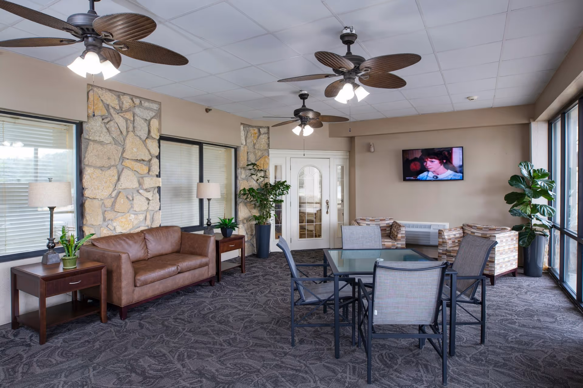 A well-lit common area with a brown leather couch flanked by two wooden side tables with lamps and plants. There are two ceiling fans with lights, a glass table with four chairs in the center, and two patterned armchairs near a wall-mounted TV. Large windows line one side of the room, and there are stone accent walls and potted plants adding to the decor.