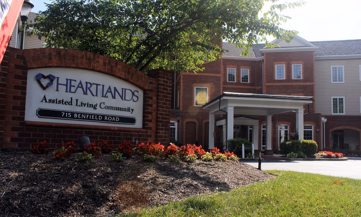 Brick entrance sign reading 'Heartlands Assisted Living Community' beside a landscaped flowerbed with the facility's brick building and porte-cochère in the background.