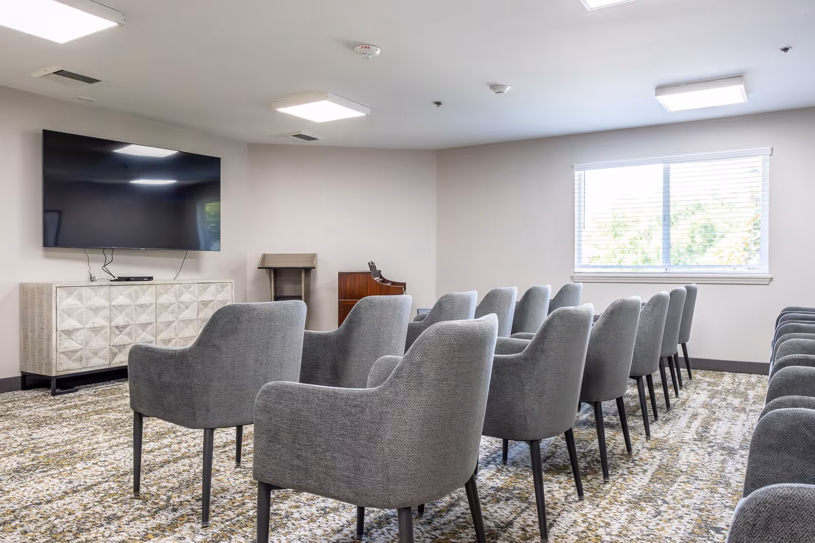 Rows of gray upholstered chairs arranged facing a wall-mounted TV in a bright meeting/media room with a window.