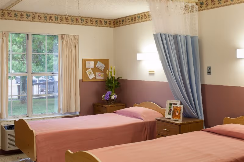 A shared bedroom in a senior living facility with two single beds covered in pink bedspreads. Each bed has a wooden headboard and a nightstand beside it. One nightstand has framed photos and the other has a vase with flowers. A blue privacy curtain is partially drawn between the beds. There is a window with beige curtains showing green trees outside. The walls are painted beige with a maroon lower half and a decorative border near the ceiling.