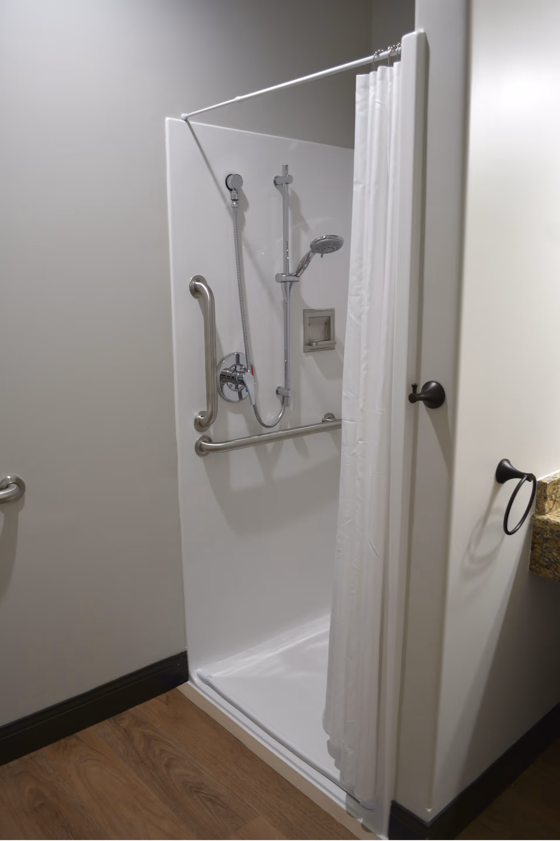 A small, accessible shower stall with a white curtain partially drawn to the side. The shower has a handheld showerhead attached to a vertical sliding bar, and two stainless steel grab bars for support. The floor outside the shower is wood, and part of a granite countertop with a towel ring is visible on the right.