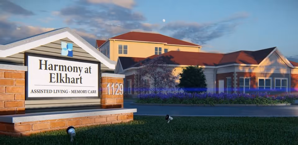 Exterior view of Harmony at Elkhart assisted living and memory care facility with a sign in the foreground displaying the facility name and address number 1129, and the building with red roofs and beige walls in the background under a partly cloudy sky.