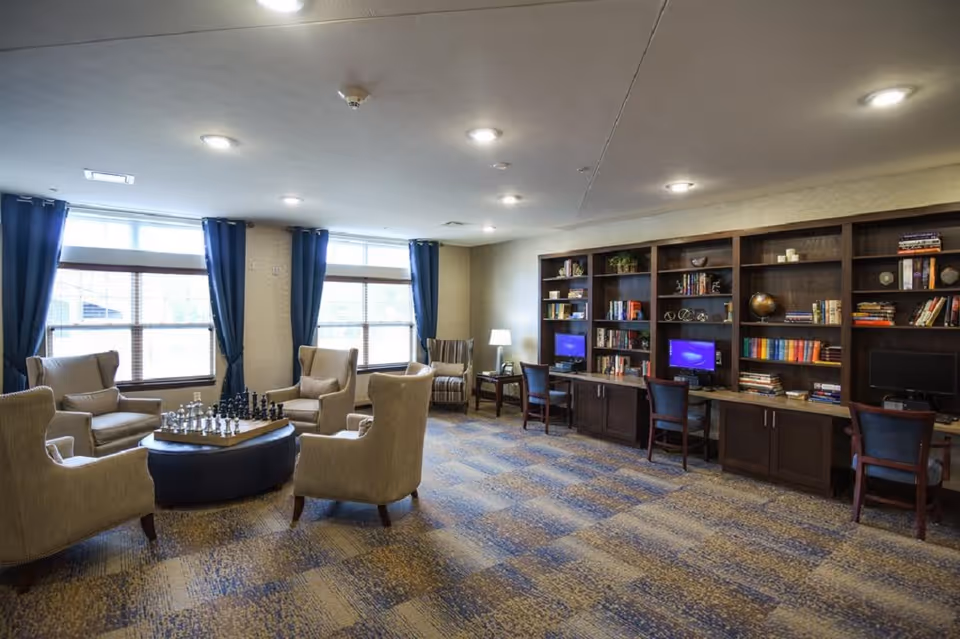 Spacious common lounge with armchairs arranged around a central table with a chess set and built-in bookshelves and computer stations along one wall.