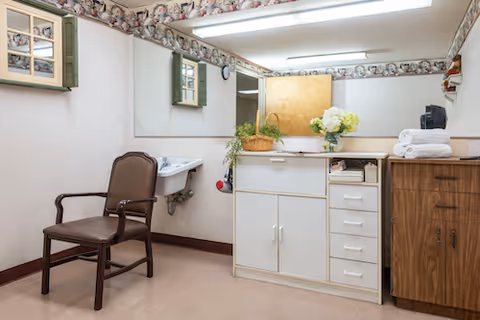 A small room with a brown chair, a wall-mounted sink, a large mirror above the sink, a white cabinet with drawers and doors, a wooden cabinet, and decorative floral wallpaper border near the ceiling. There is a basket with greenery and a vase with white flowers on the white cabinet, along with neatly folded white towels on the wooden cabinet.