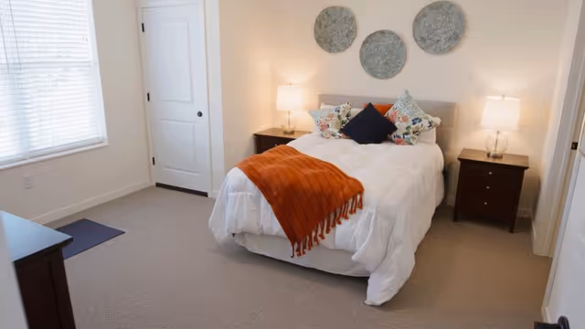 Bright bedroom with a bed, orange throw, pillows, two nightstands, and lamps.