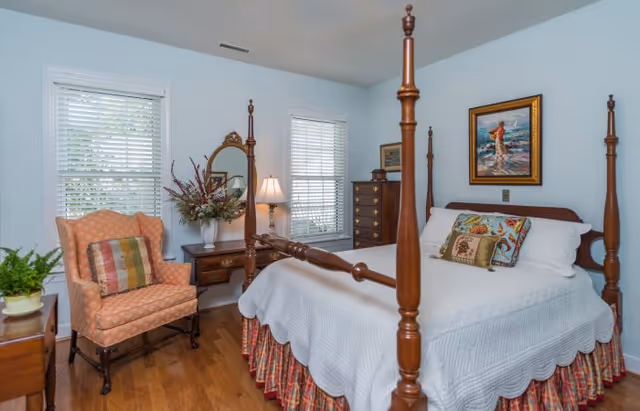 Light, traditionally furnished bedroom with a four-poster wooden bed, bedside tables, an upholstered armchair, and windows with blinds.