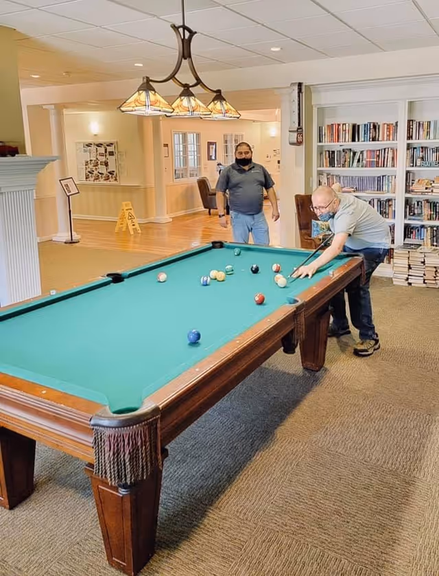 Two men playing pool in a well-lit room with a green pool table. One man is leaning over the table aiming a shot, while the other stands nearby watching. The room has bookshelves filled with books, a carpeted floor, and a hanging light fixture above the pool table.