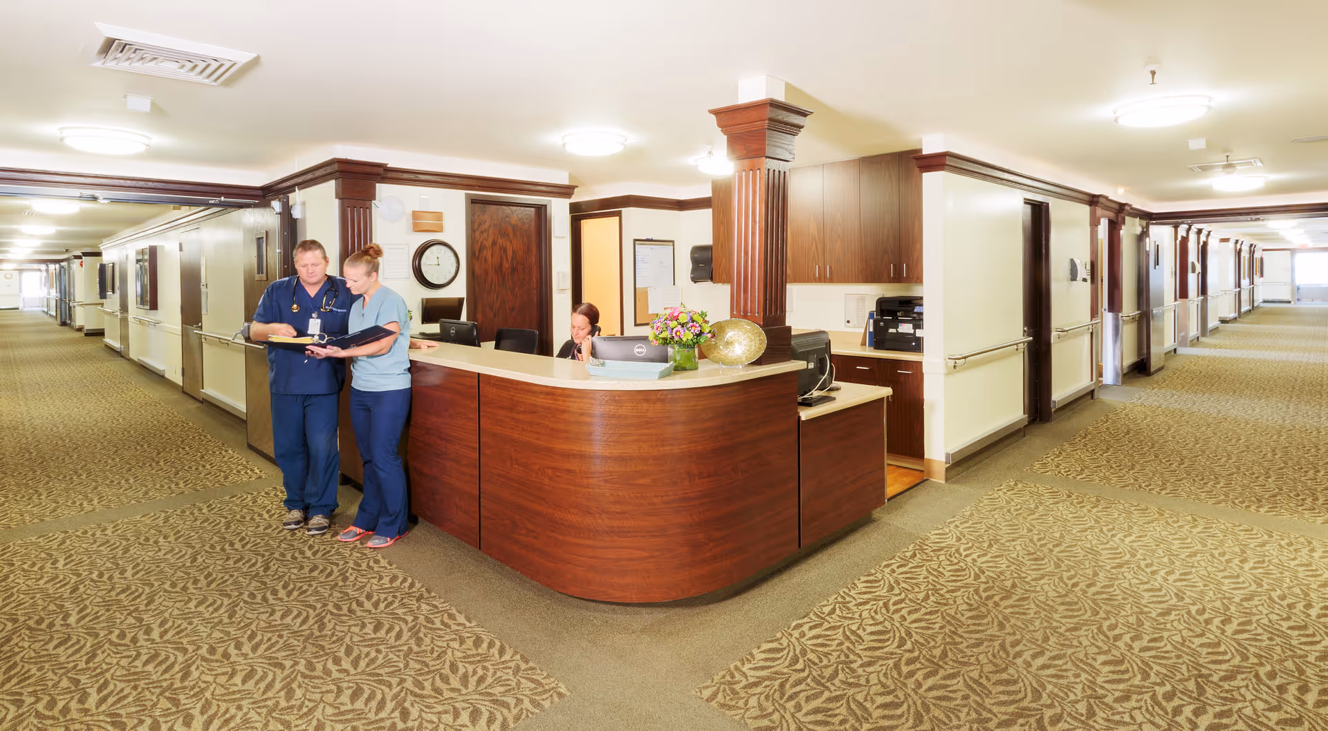 A wide, well-lit hallway in a senior living facility with a wooden reception desk at the center. Two healthcare workers in scrubs are standing and discussing something on a clipboard, while another staff member is seated behind the desk working on a computer. The hallway has patterned carpet, handrails along the walls, and multiple doors leading to rooms.