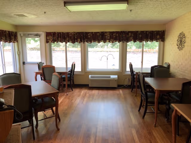 Sunlit communal dining room with several wooden tables and chairs, hardwood floors, and large windows with a wall-mounted air unit beneath them.