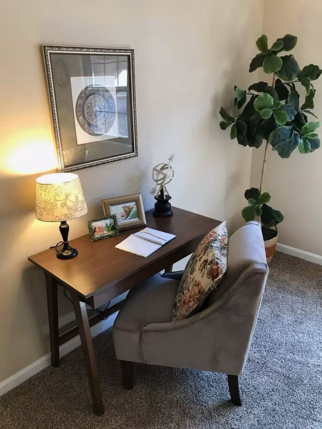 A cozy corner with a wooden desk and a cushioned chair featuring a floral pillow. On the desk, there is a decorative lamp, two framed photos, an armillary sphere, and an open notebook with a pen. A large green potted plant stands next to the desk against a beige wall with a framed circular artwork hanging above.