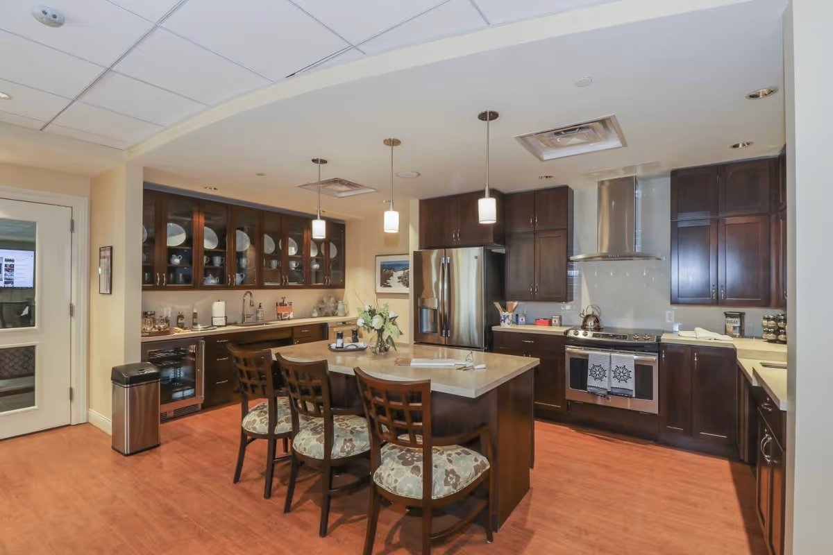 A modern kitchen with dark wood cabinets, a stainless steel refrigerator, stove with a range hood, and a central island with three cushioned chairs. The kitchen has pendant lights hanging from the ceiling and glass-front cabinets displaying dishes. The floor is wooden, and there is a trash bin near a door with glass panels.