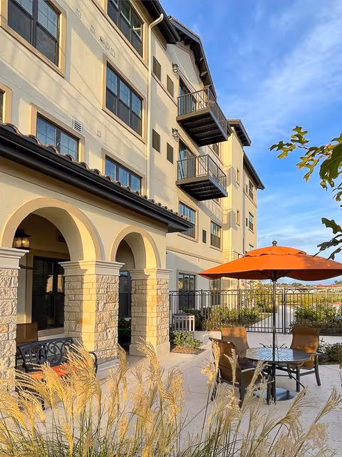 Outdoor patio area of a multi-story senior living facility with beige walls and balconies. The patio features a round table with four chairs and an orange umbrella. Ornamental grasses and plants surround the patio, and the sky is clear and blue.