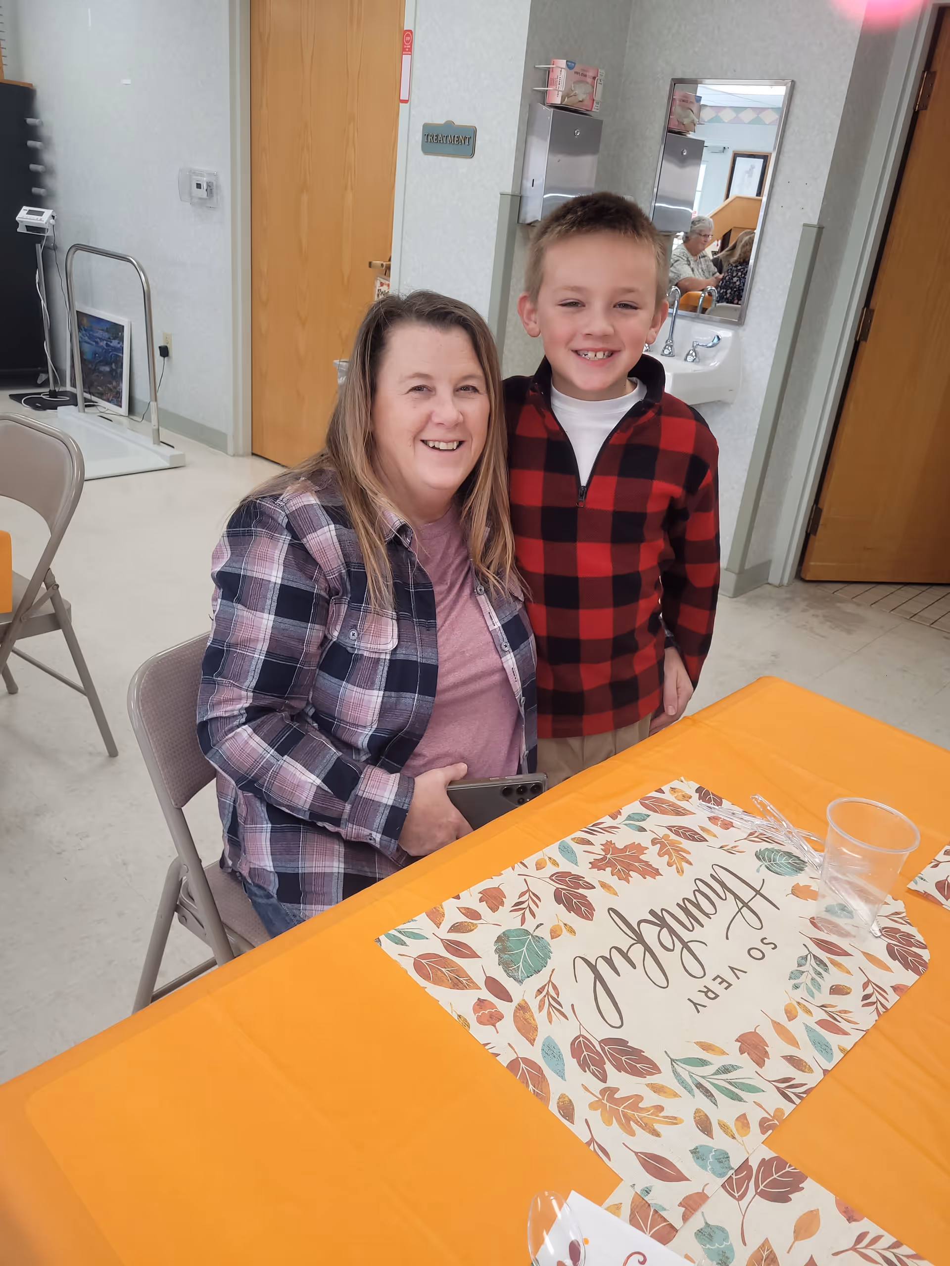 A woman and a young boy smiling and posing together at a table covered with an orange tablecloth and a decorative placemat with autumn leaves and the words 'SO VERY thankful'. They are indoors in a room with beige walls, a wooden door, a sink, and a mirror in the background.