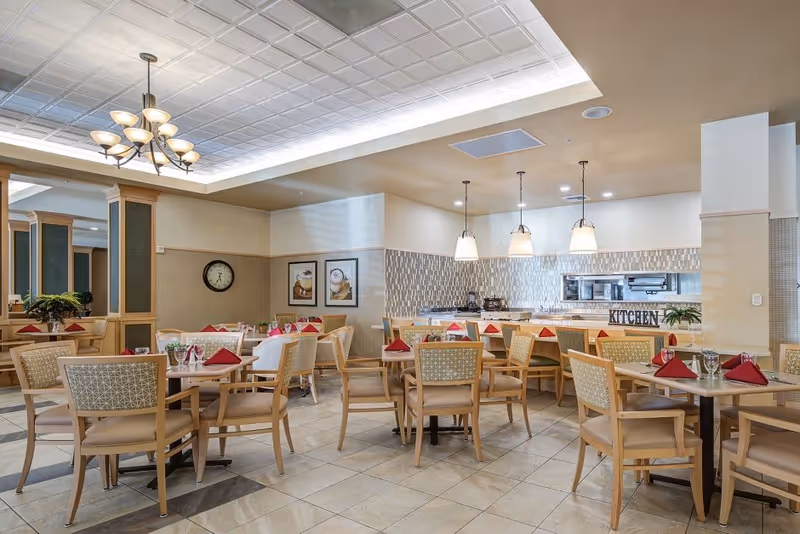 A bright dining room in a senior living facility with multiple wooden tables and chairs arranged neatly. Each table is set with red folded napkins, glasses, and small flower vases. The room features a tiled floor, a decorative ceiling with recessed lighting, and a chandelier. In the background, there is a kitchen area with pendant lights hanging above the counter and the word 'KITCHEN' displayed on the counter. The walls have framed artwork and a clock.