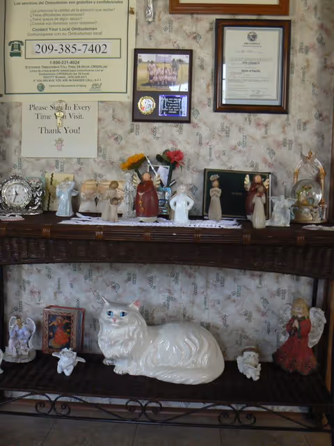 Console table against floral wallpaper displaying porcelain figurines and a large white ceramic cat beneath framed certificates.