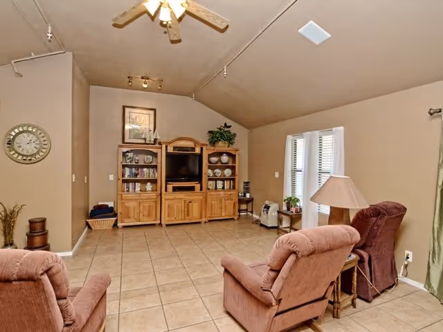 A cozy living room with beige walls and tiled floor featuring two recliner chairs facing a wooden entertainment center with a TV, books, and decorative items. There is a ceiling fan above, a wall clock on the left, and windows with white blinds on the right letting in natural light.