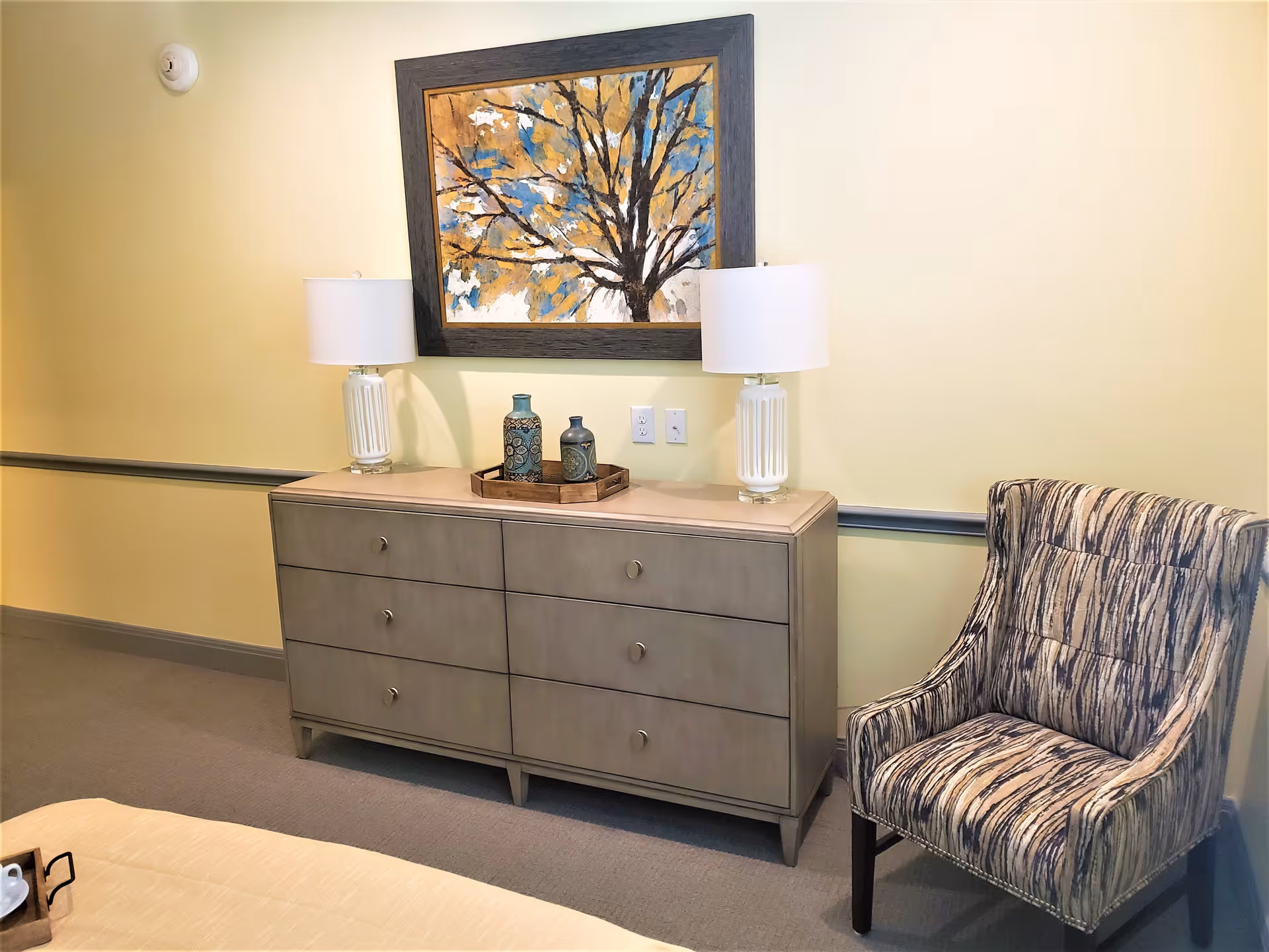 A cozy interior corner featuring a gray six-drawer dresser with two white table lamps and decorative vases on top, a framed painting of a tree with yellow and blue leaves hanging above, and a patterned upholstered armchair to the right.