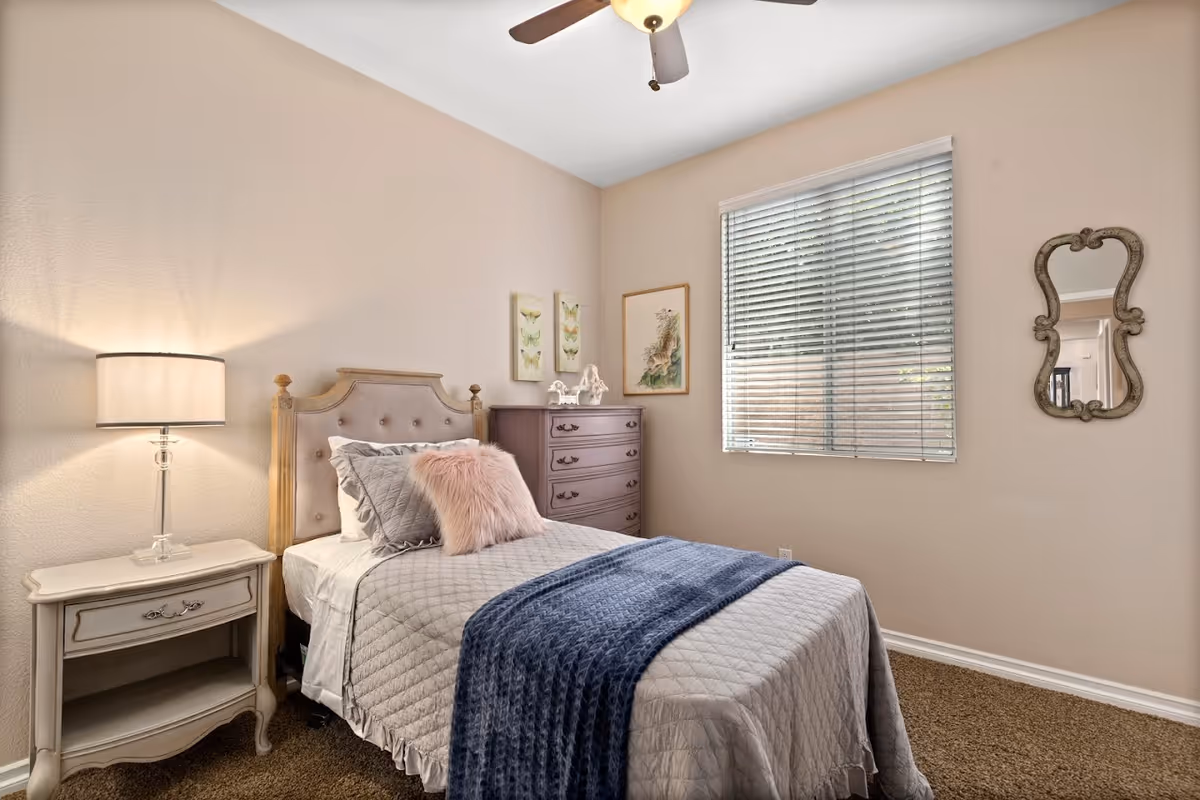 A cozy bedroom with a single bed featuring a tufted headboard, gray quilted bedding, a pink fluffy pillow, and a blue throw blanket. Next to the bed is a white nightstand with a lamp, and against the wall is a wooden dresser with decorative items on top. The room has beige walls, a window with blinds, a ceiling fan, and a decorative mirror on the wall.