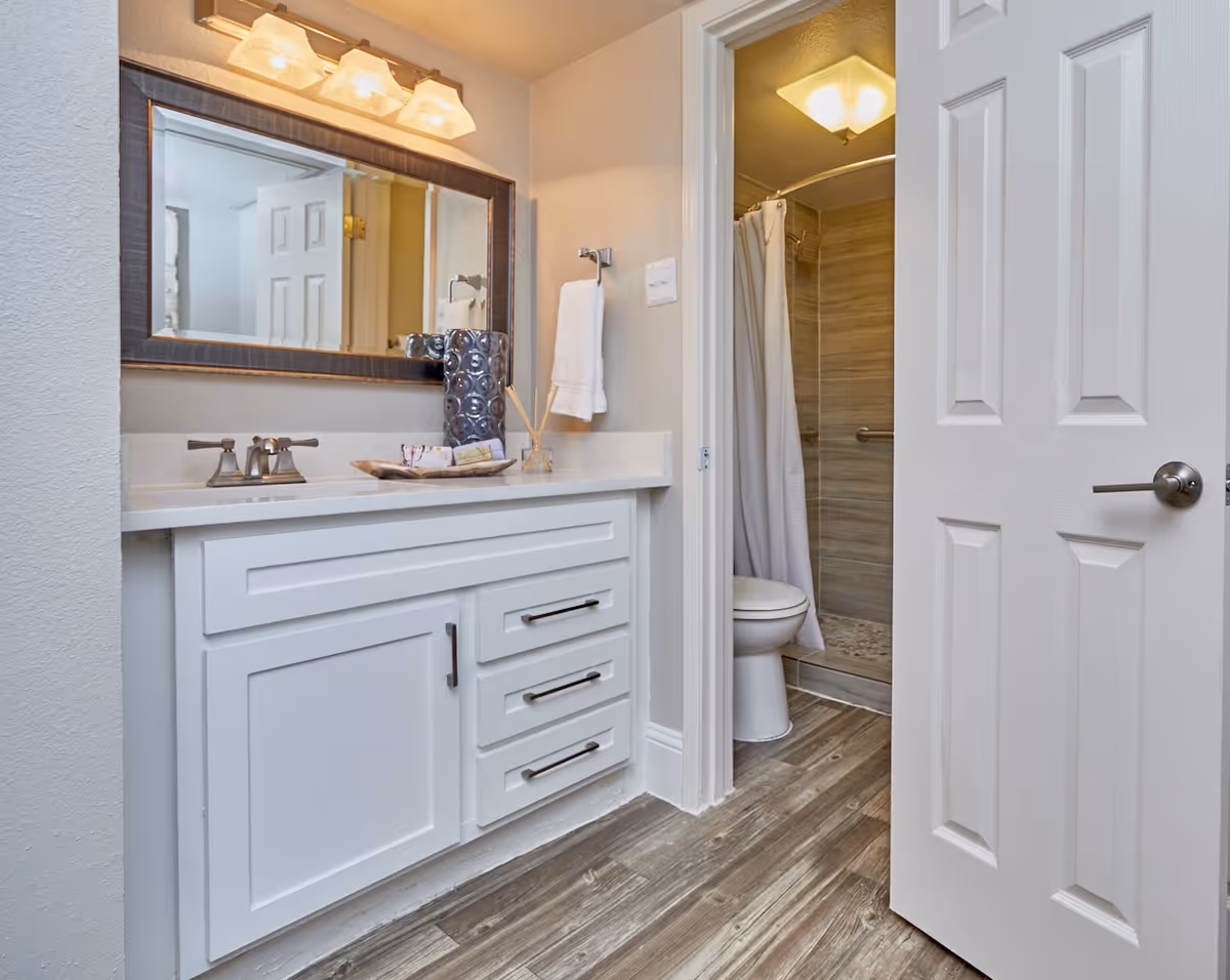 A clean and modern bathroom featuring a white vanity with drawers and a large mirror above it. On the countertop, there is a decorative vase, a tray with soap, and a reed diffuser. To the right, an open door reveals a toilet and a shower area with a curtain and tiled walls. The floor has wood-like tiles.