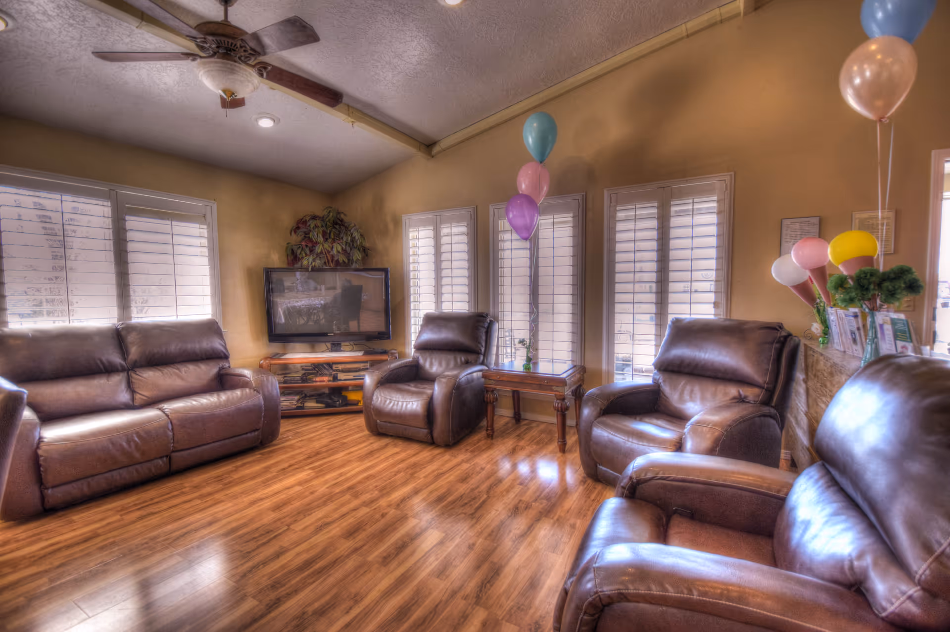 A cozy living room with brown leather sofas and armchairs arranged around a flat-screen TV mounted on a wooden stand. The room has wooden flooring, beige walls, and three windows with white shutters. There are colorful balloons tied to a small wooden table and a counter, adding a festive touch. A ceiling fan with lights is mounted on the ceiling.